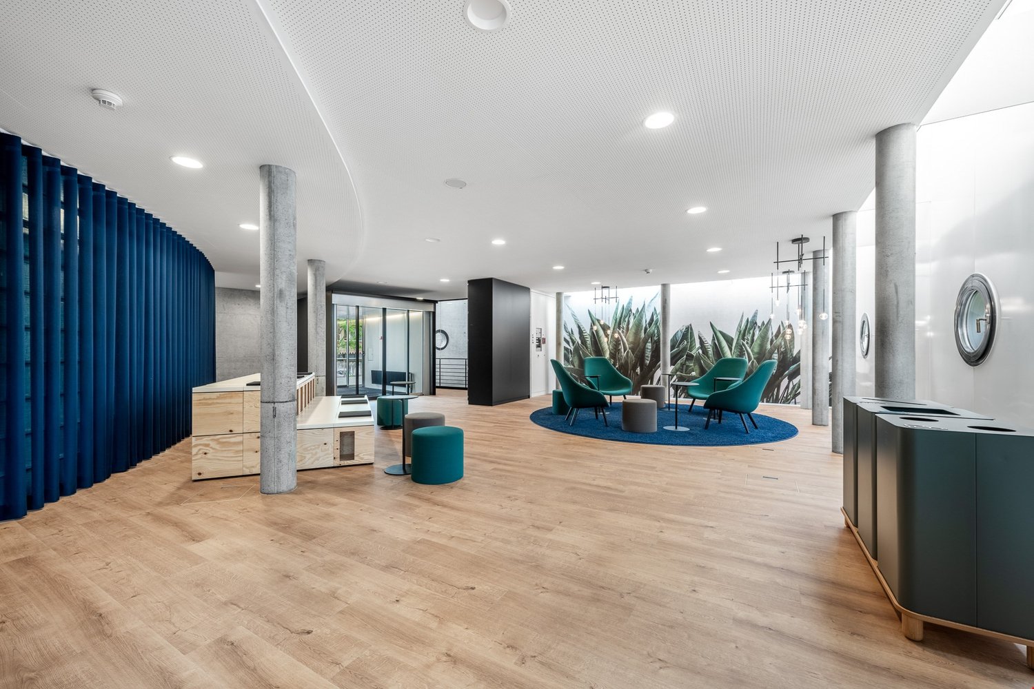 Spacious lobby with hardwood floors, white walls, blue curtains, modern furniture including green armchairs and ottomans, large wall mural with tropical plants, and various storage units.