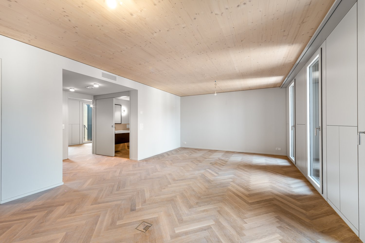 Empty room with wooden parquet flooring, white walls, and a wooden ceiling. There are two doors, one leading to a bathroom and the other to another room.