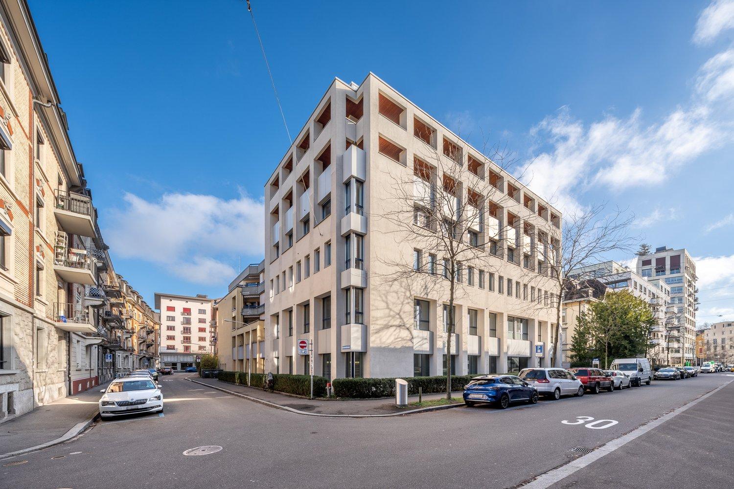 Modern multi-story building with glass windows, balconies, and a parking area in front.