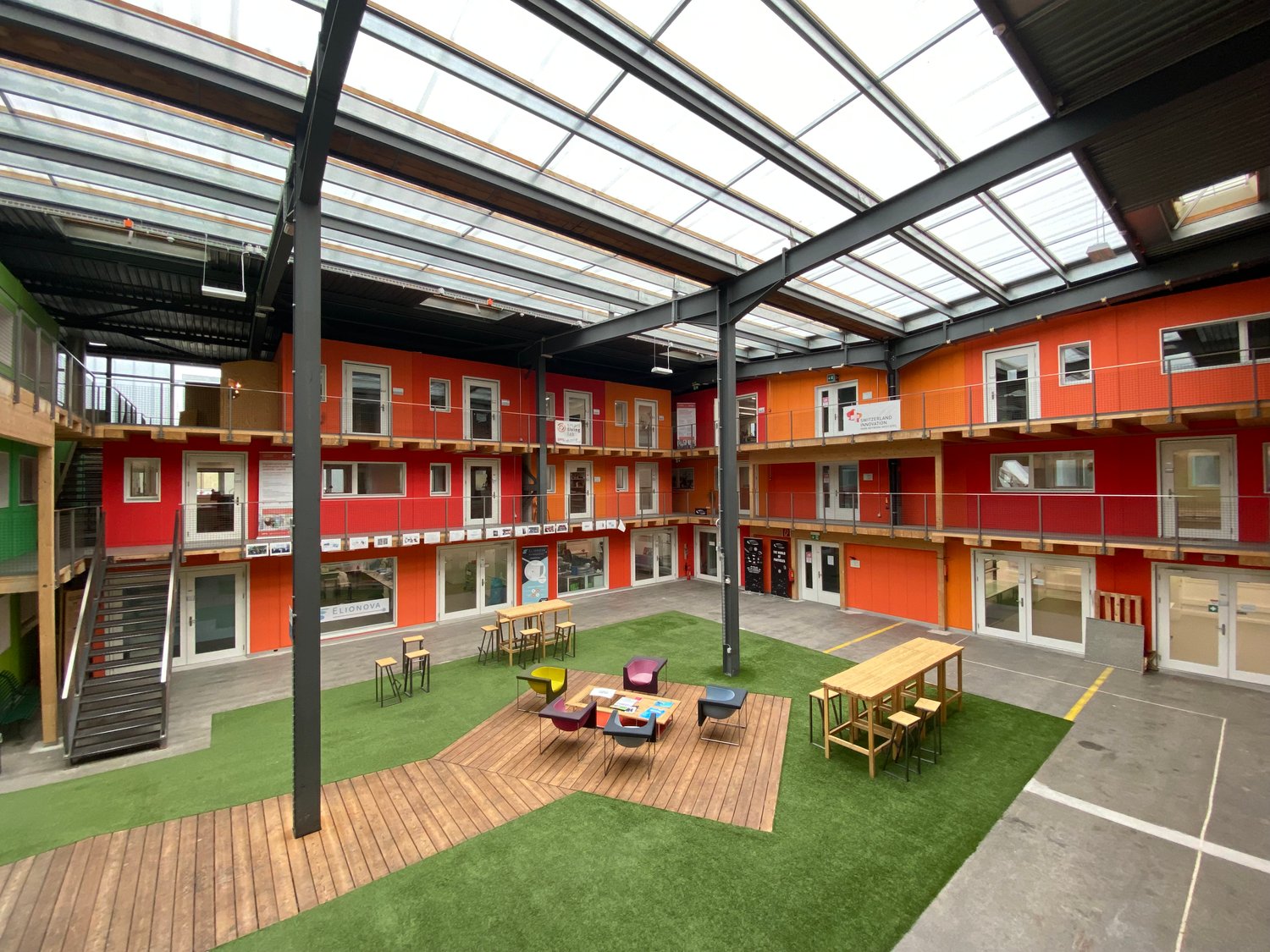 Large open interior space with a grass lawn, wooden seating area, tables, and staircases; orange and red walls with glass doors and windows.