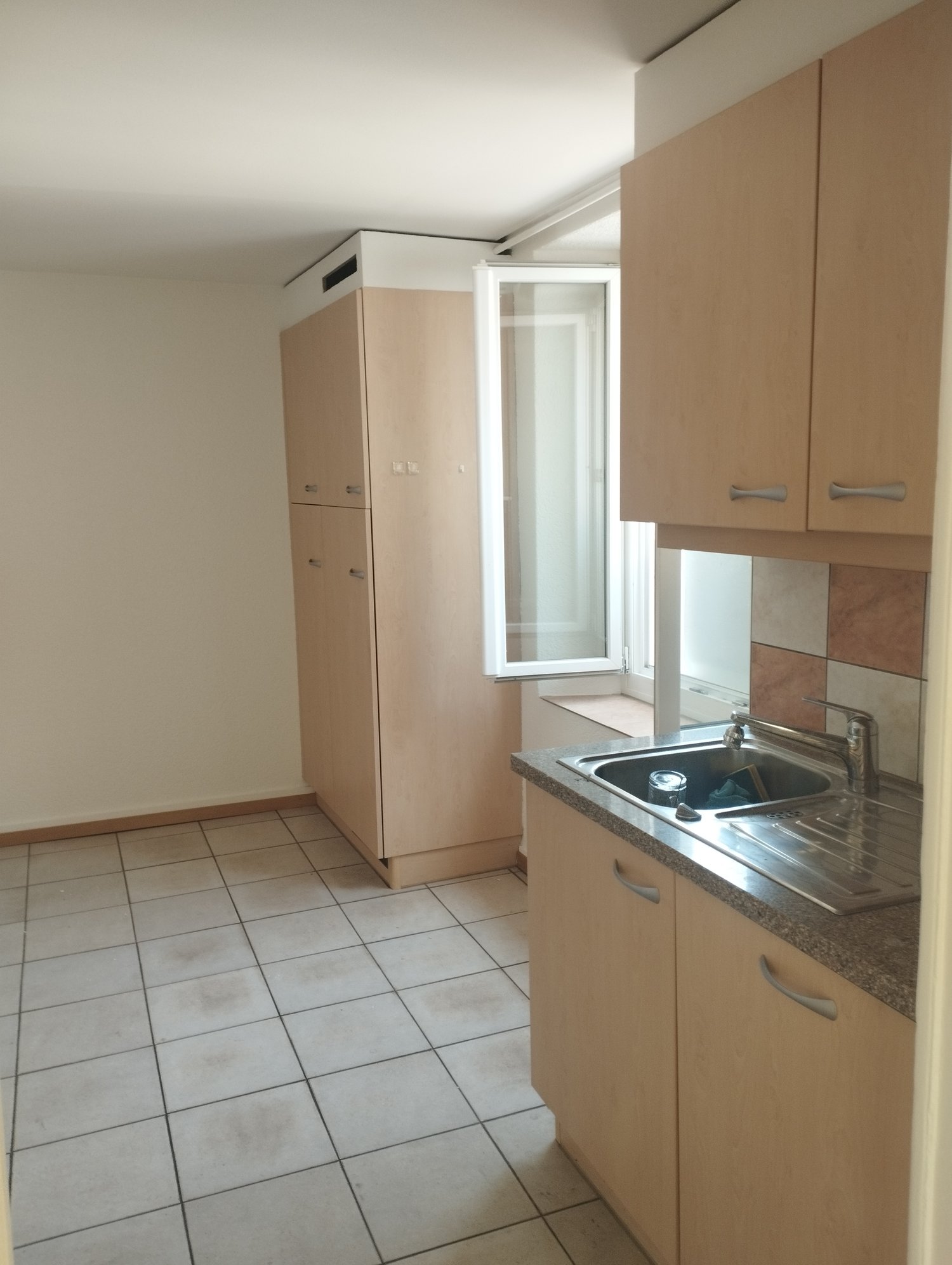 The image shows a kitchen area with beige cabinets, a sink, and a refrigerator. The floor is tiled, and there is a window visible. The kitchen appears to be part of a larger living space.
