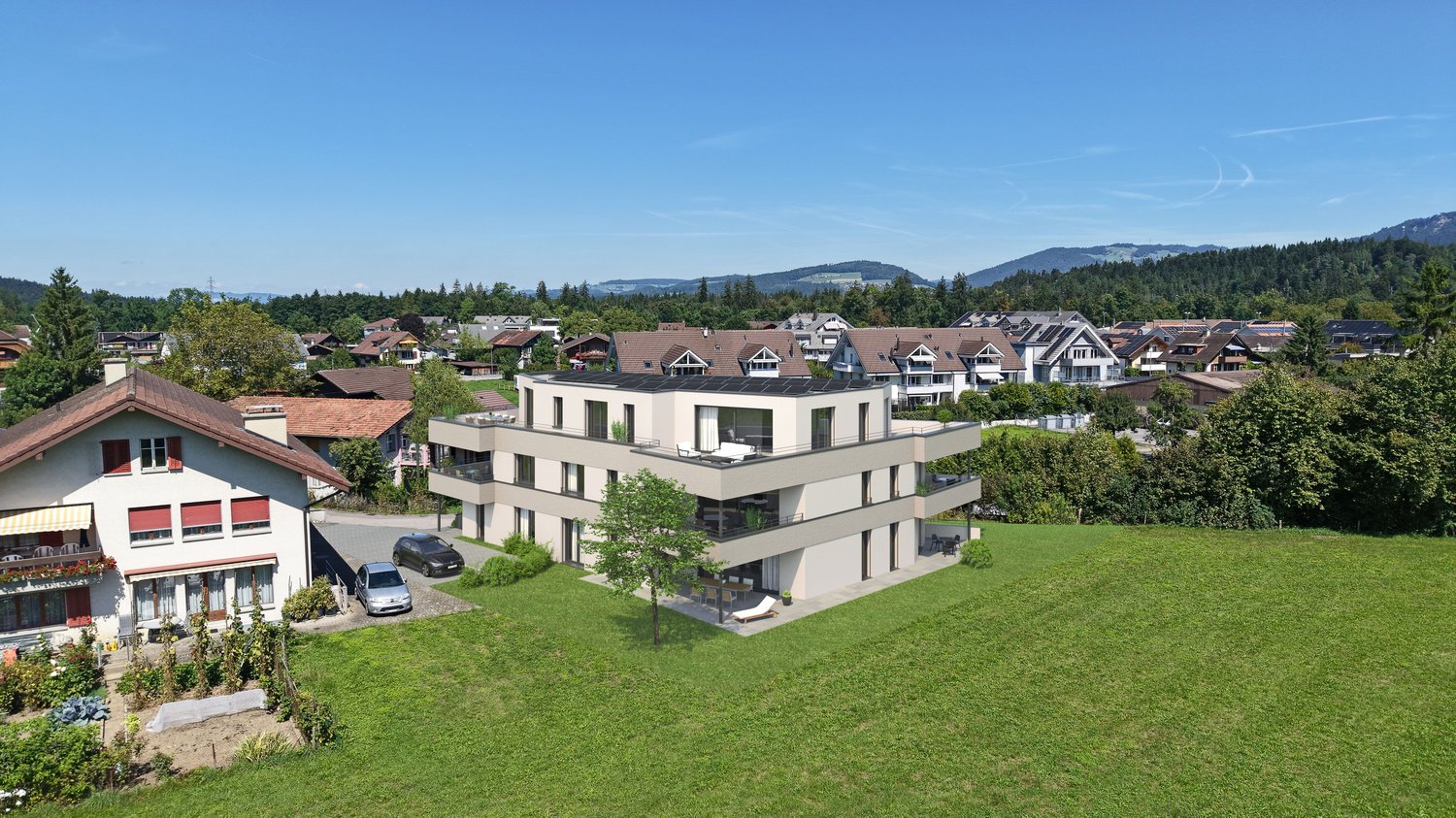 Aerial view of a modern building with balconies, located in a village with several houses and a beautiful mountain landscape in the background.