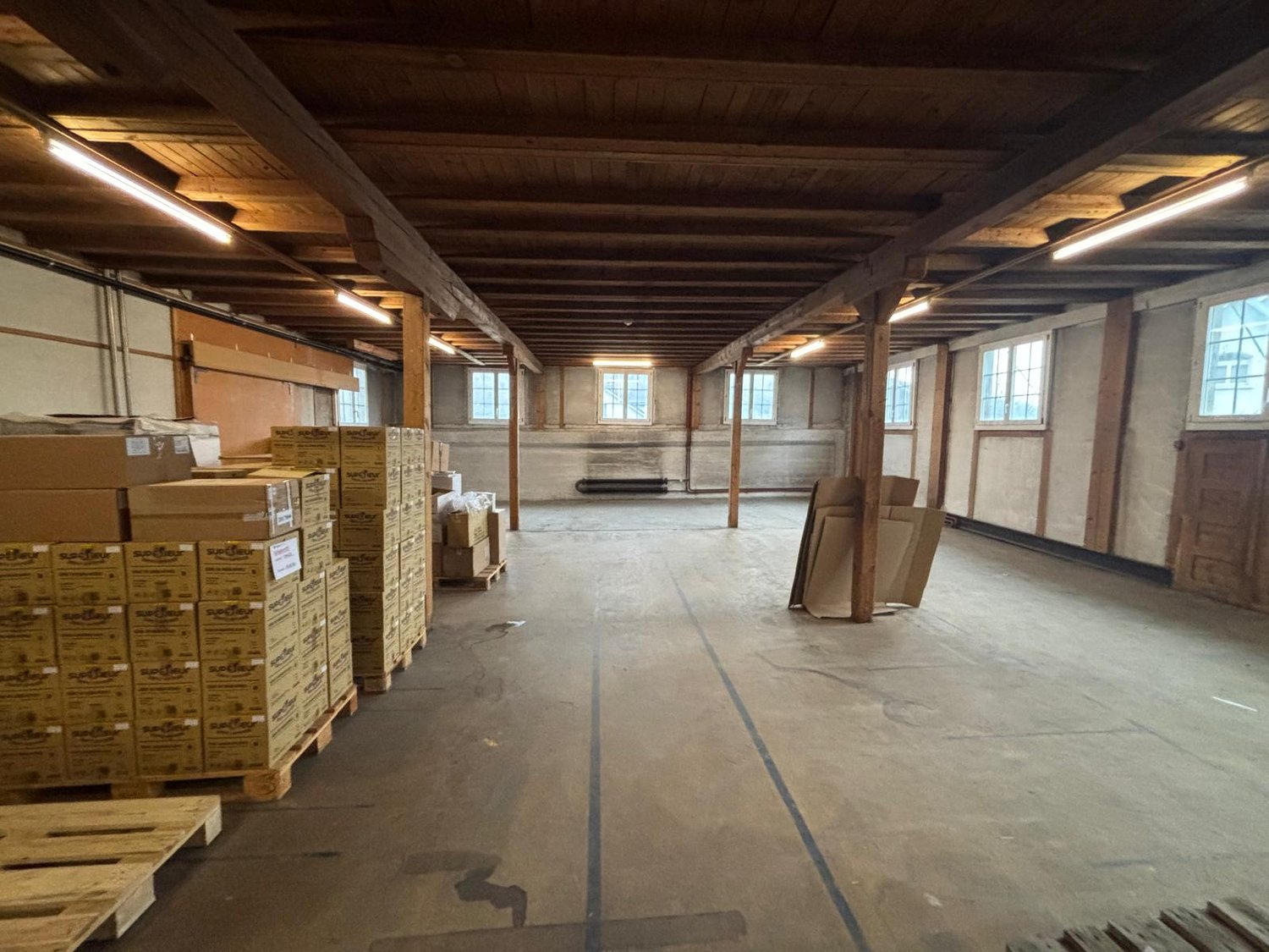 Empty warehouse with wooden beams, fluorescent lights, wooden pallets, stacks of boxes, concrete floor, windows
