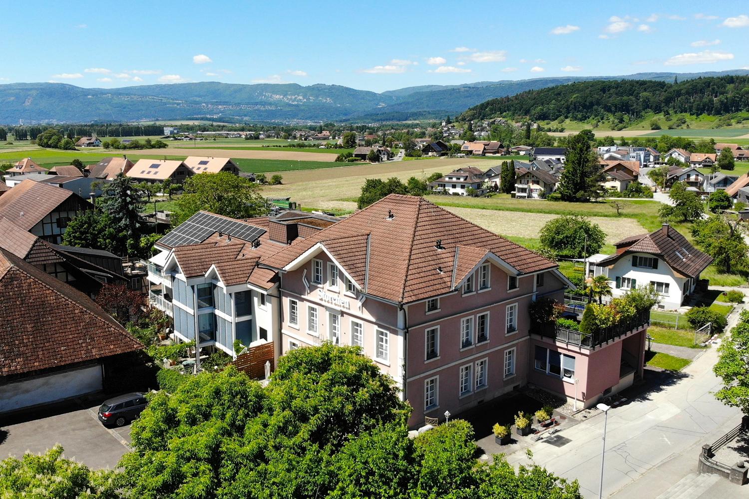 The image shows a scenic rural town with a mix of residential and commercial buildings. The buildings have red tile roofs and are surrounded by lush greenery, including trees and fields. There are several multi-story apartment buildings with balconies vis