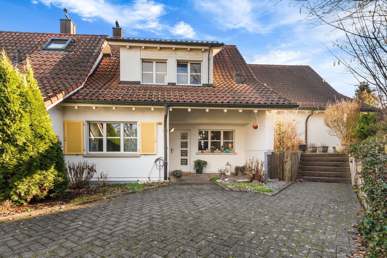 Detached 2-story house, red tiled roof, white walls, yellow shutters, a driveway, fenced front yard, several plants in front