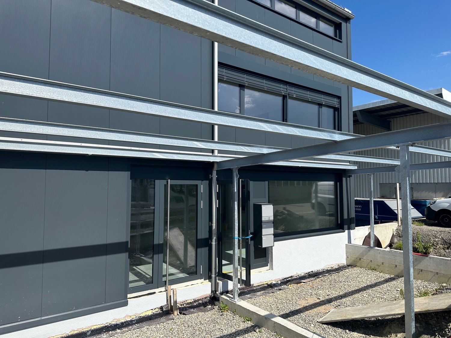 Two-story commercial building with a modern, minimalist design. The exterior is made of dark gray panels and features large windows. There is a covered entrance area with a glass door. The building appears to have a parking area in front.