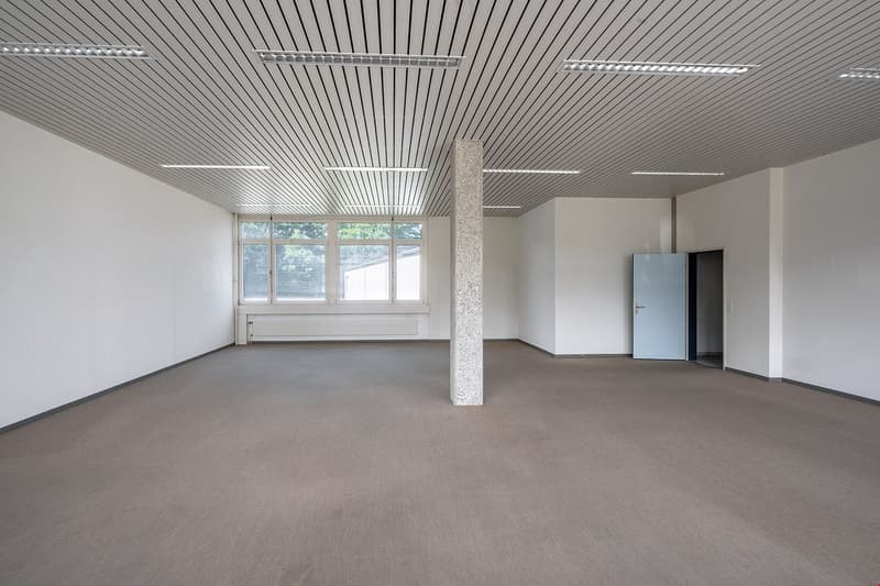 Large open office space with high ceilings, exposed beams, and large windows. The room has a concrete column in the center and a refrigerator in the corner. The space appears to be unfinished and ready for customization.