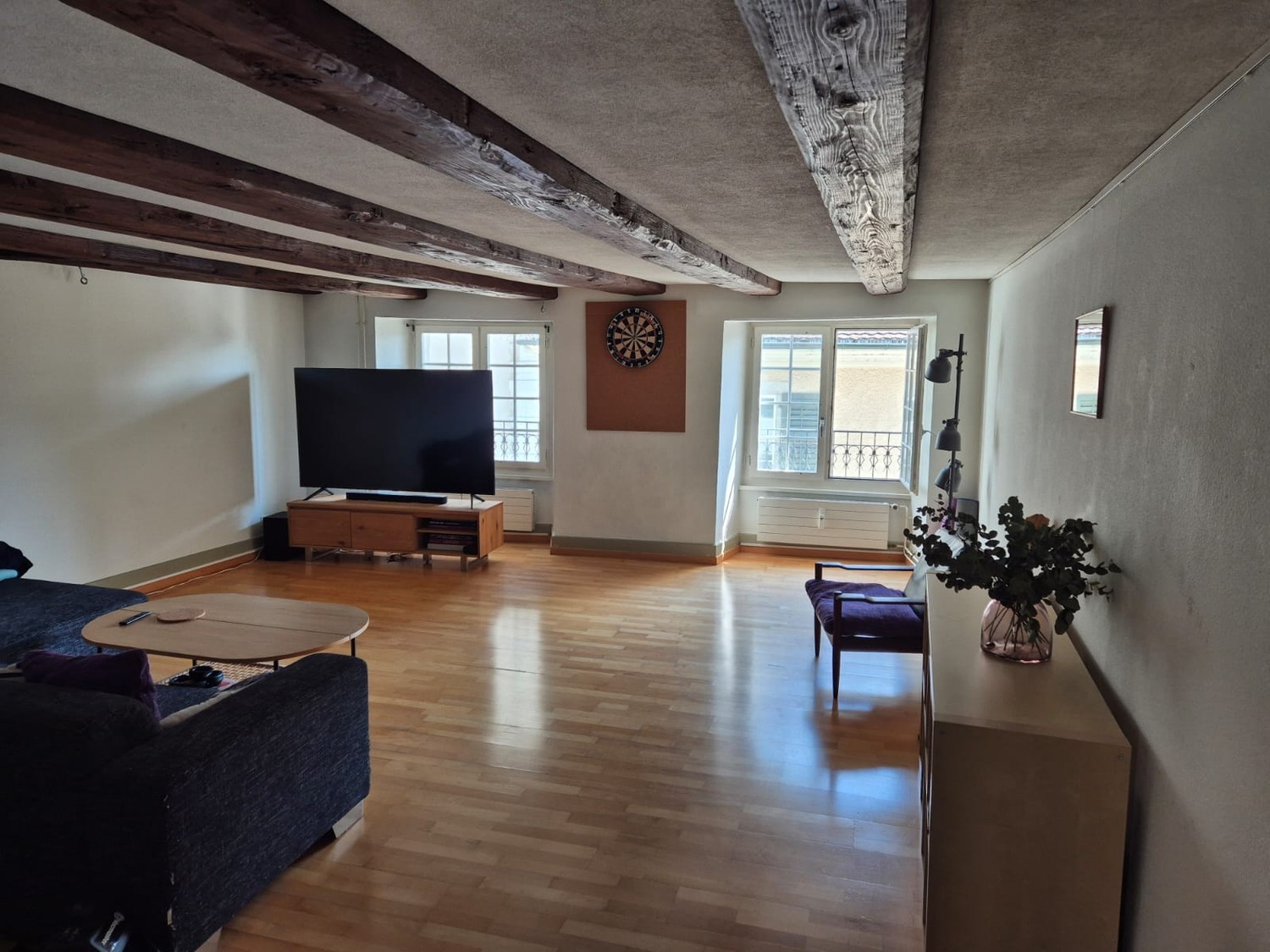 Spacious living room with hardwood floors, exposed wooden beams on the ceiling, a large TV mounted on the wall, a sofa, and a coffee table. There is also a dart board on the wall and a potted plant in the corner.