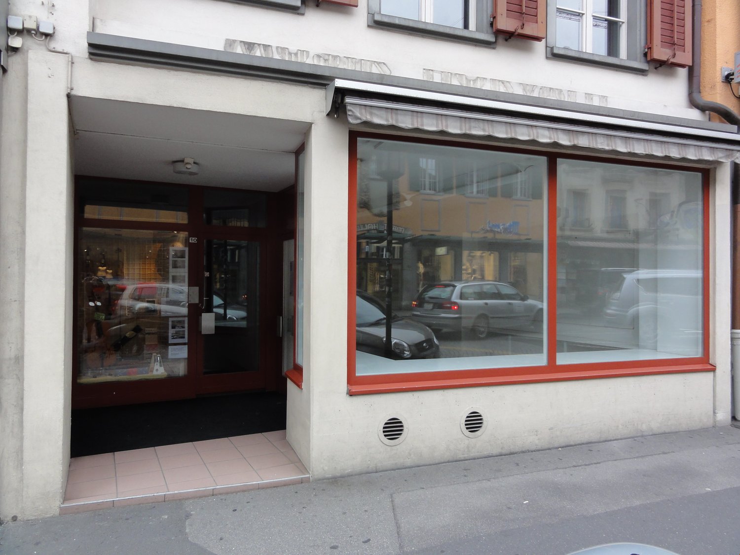 The image shows the exterior of a commercial building with a storefront. The building has large windows with red frames, and the entrance is through a doorway. There are cars parked on the street in front of the building.