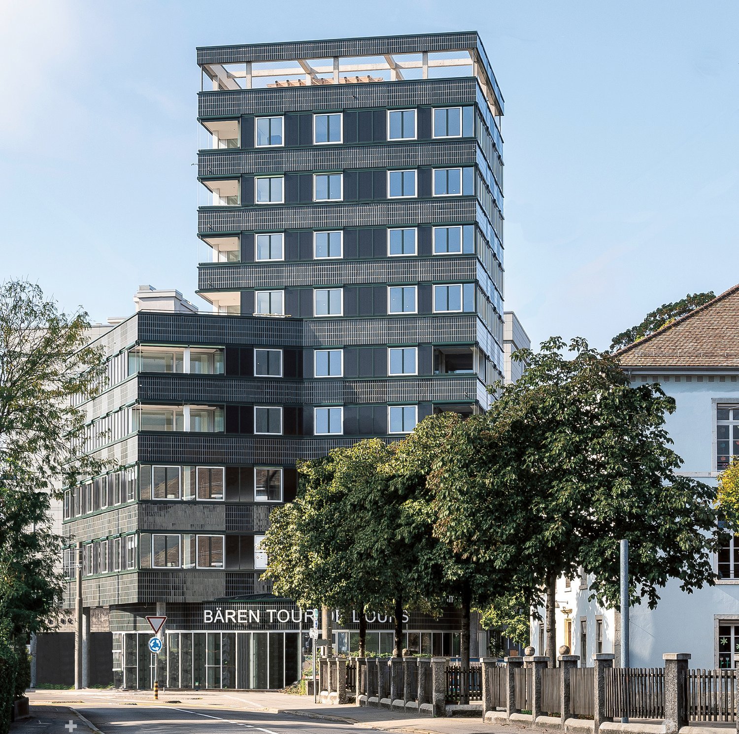 Multi-story commercial building with black and glass facade, balconies, and trees in the foreground