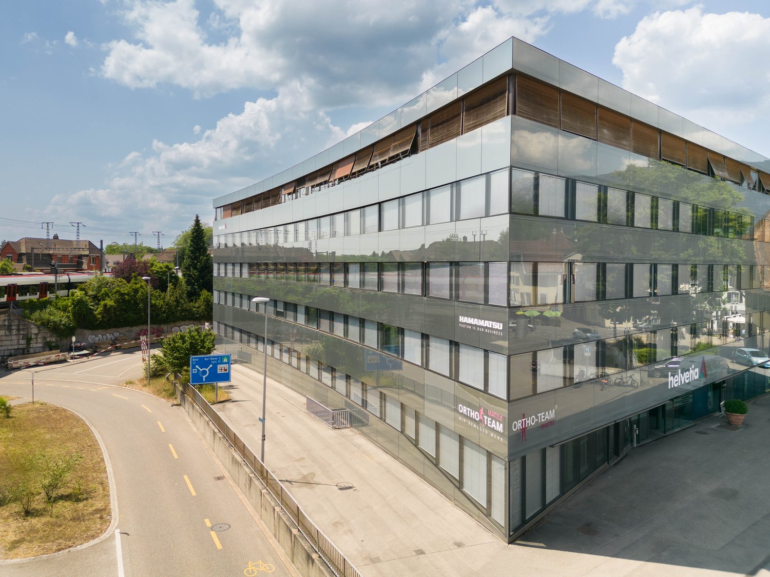 Modern office building with glass facade, multiple floors, reflective windows, outdoor parking area, street sign, greenery, and a road