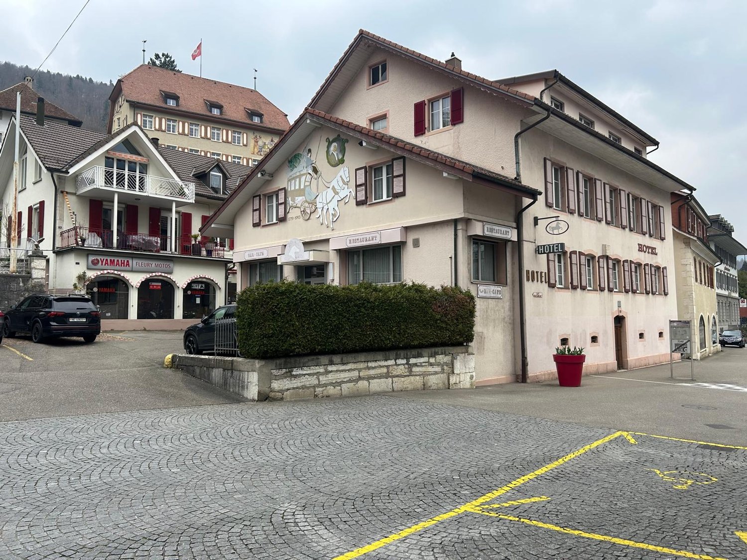 A multi-story building with a Yamaha motorcycle dealership on the ground floor, surrounded by other buildings in a small town or village setting. The building has a traditional Swiss-style architecture with red-tiled roofs and decorative elements on the f