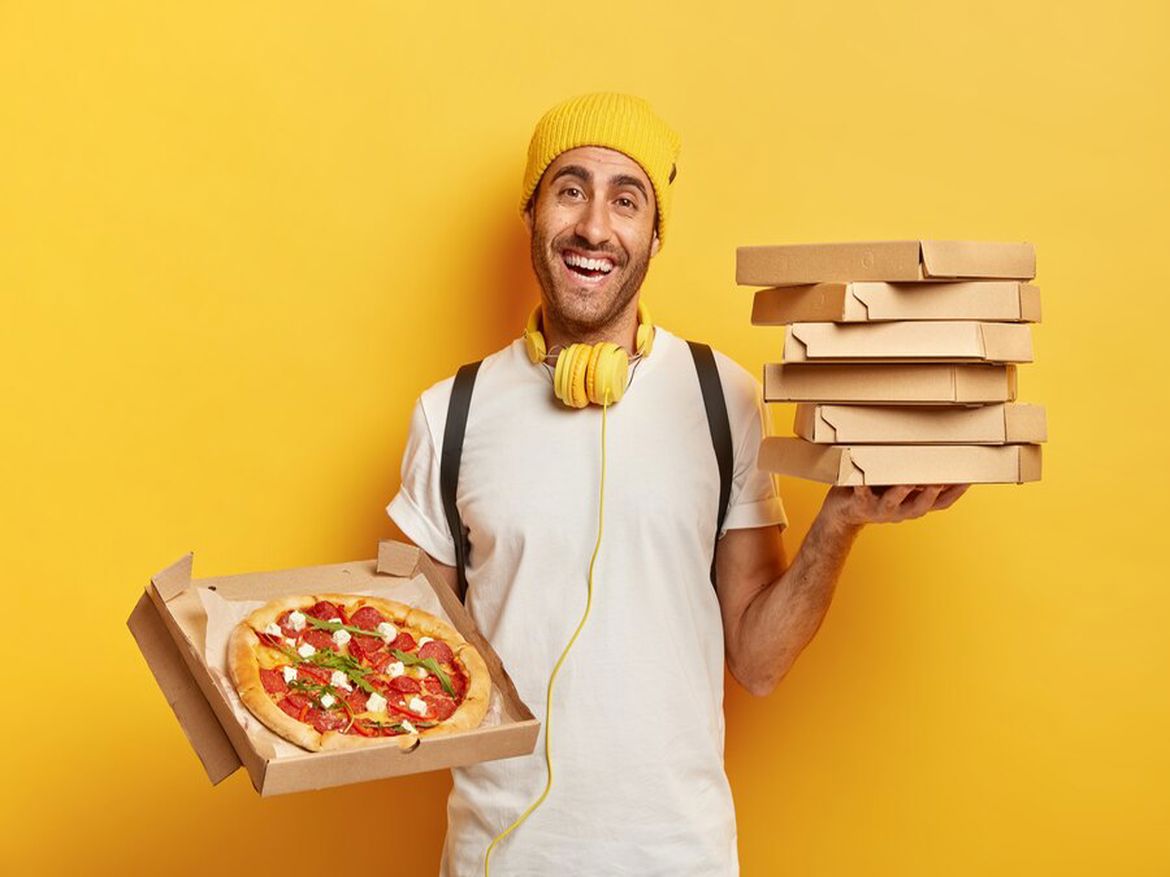 Man holding pizza boxes, smiling, wearing yellow headphones and a yellow hat