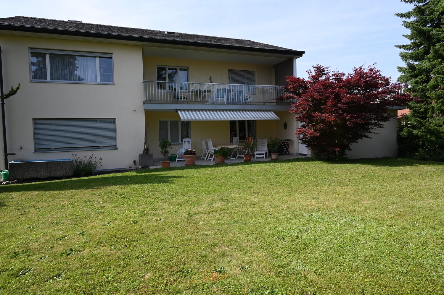 2-story house, beige exterior, terrace on the second floor, lawn, trees, plants, awning over the terrace