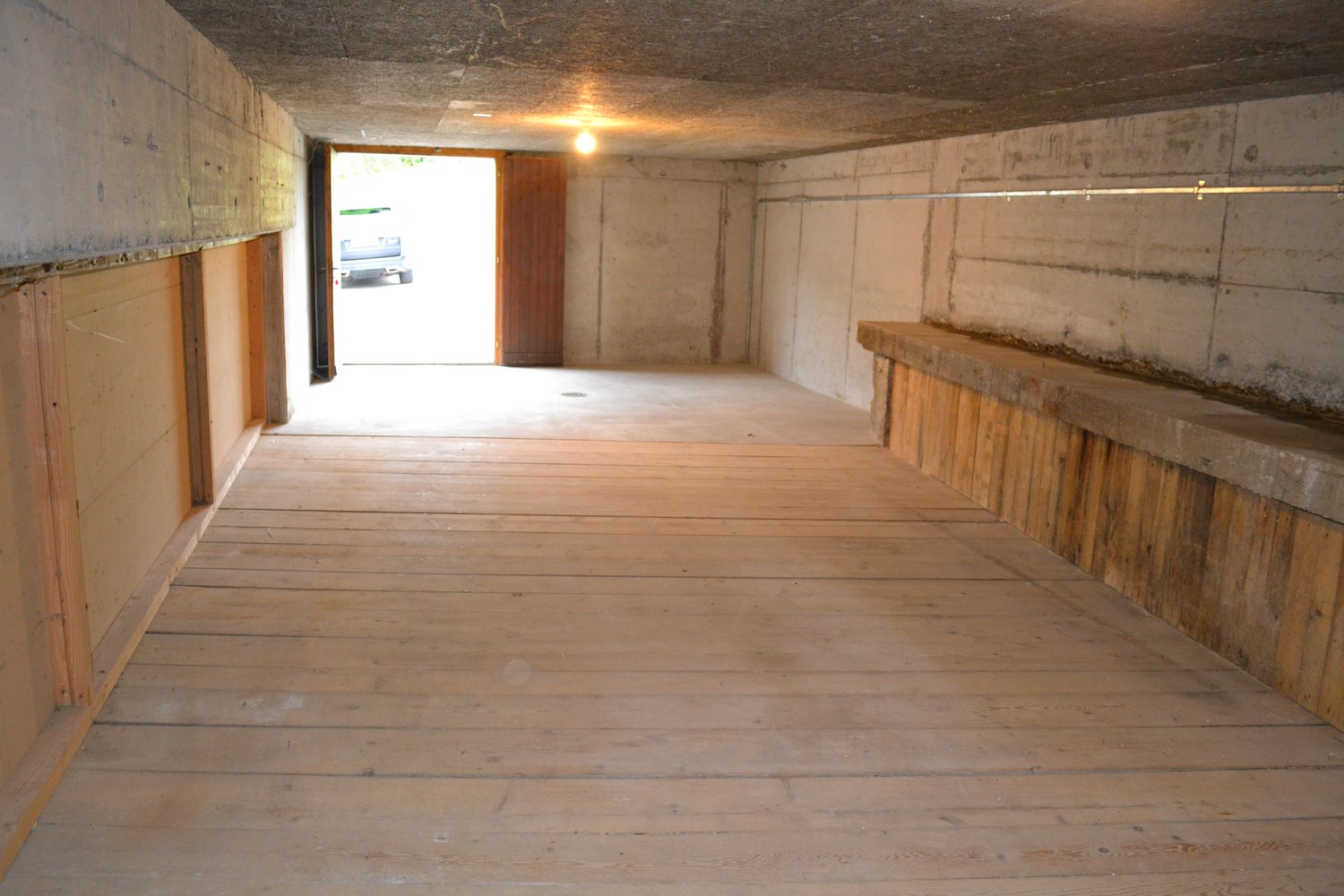 This image shows the interior of a garage or storage space. The space has concrete walls and a wooden floor. There is a window at the end of the room, allowing natural light to enter. The room appears to be empty and unfinished, with a simple light fixtur