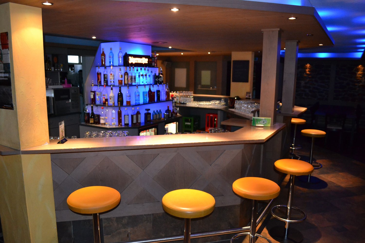 Modern bar with a sleek, geometric design. The bar counter is made of wood and has a granite-like surface. The back bar is well-stocked with various liquor bottles and glassware. There are yellow bar stools in front of the counter, and the lighting create