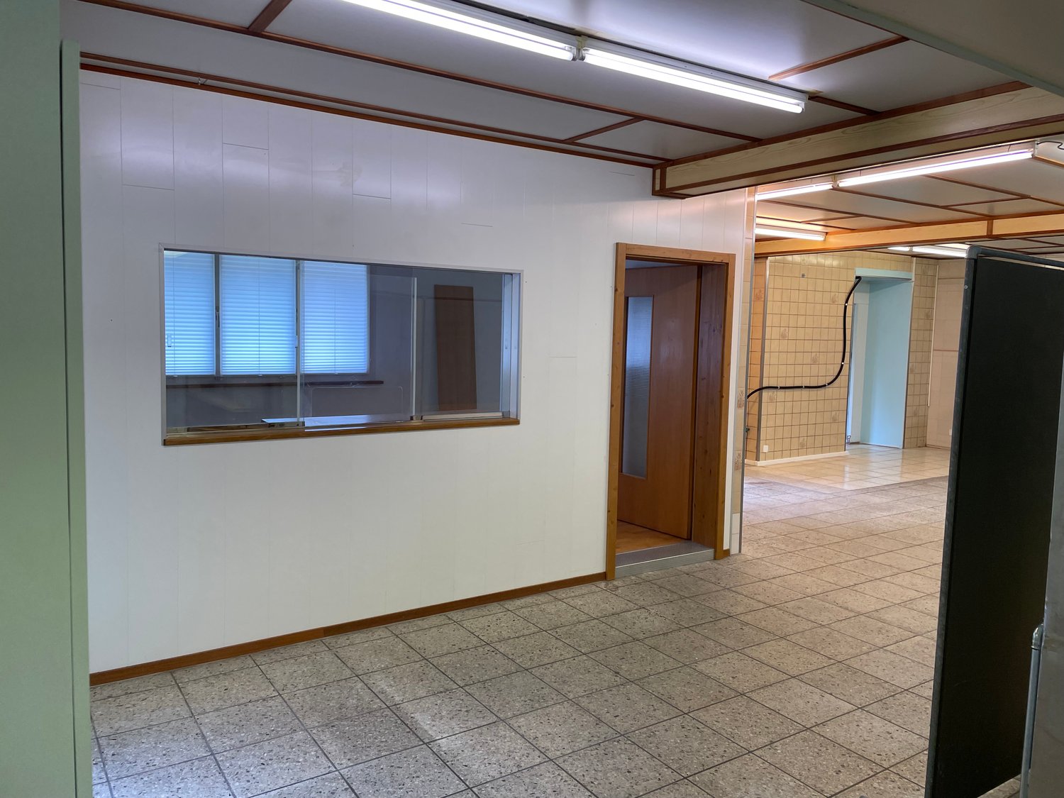 The image shows a large open office space with a tiled floor, white walls, and wooden beams on the ceiling. There is a window with blinds and a doorway leading to another room. The space appears to be a commercial or office setting.