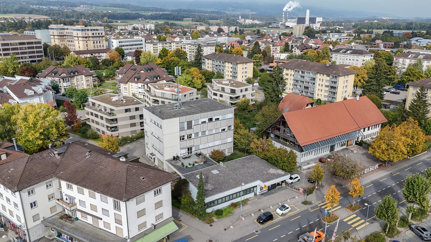 Aerial view of several multi-story residential buildings, green spaces, and trees. Several vehicles parked in the area.