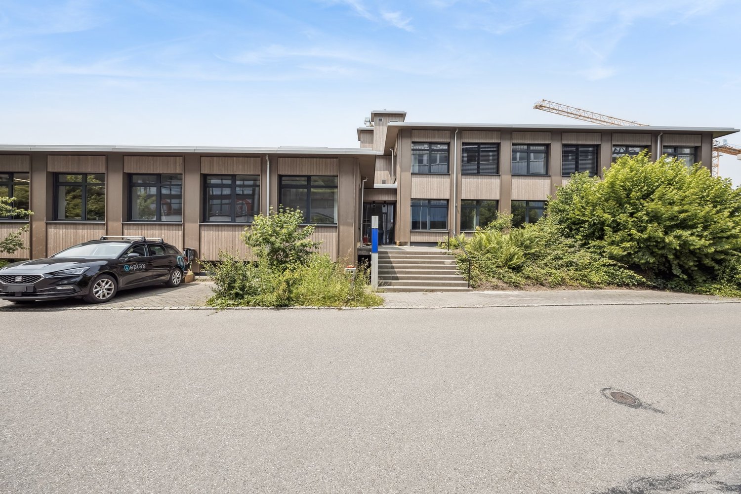 2-story commercial building with a modern, wooden exterior. The building has large windows and a covered entrance with stairs leading up to the main entrance. There is a parked car in the foreground, indicating a parking area or lot nearby.