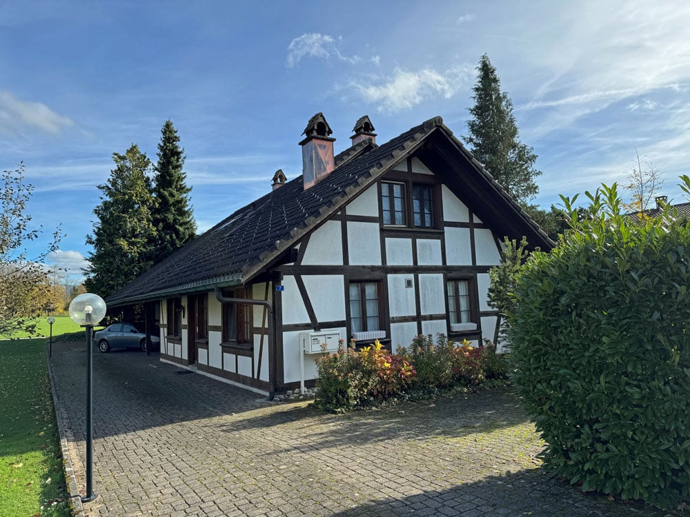 Detached house, black roof, white and brown walls, chimneys, car parked in front, brick driveway, garden, trees