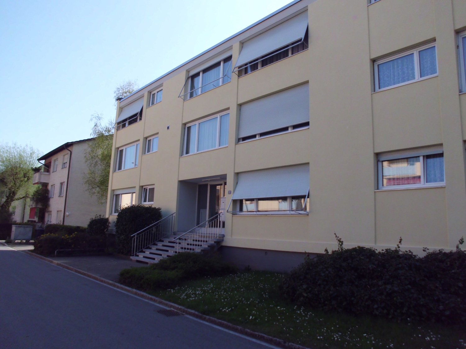 A yellow building with multiple windows and a staircase leading to the entrance. The building has a flat roof and is surrounded by bushes and grass.