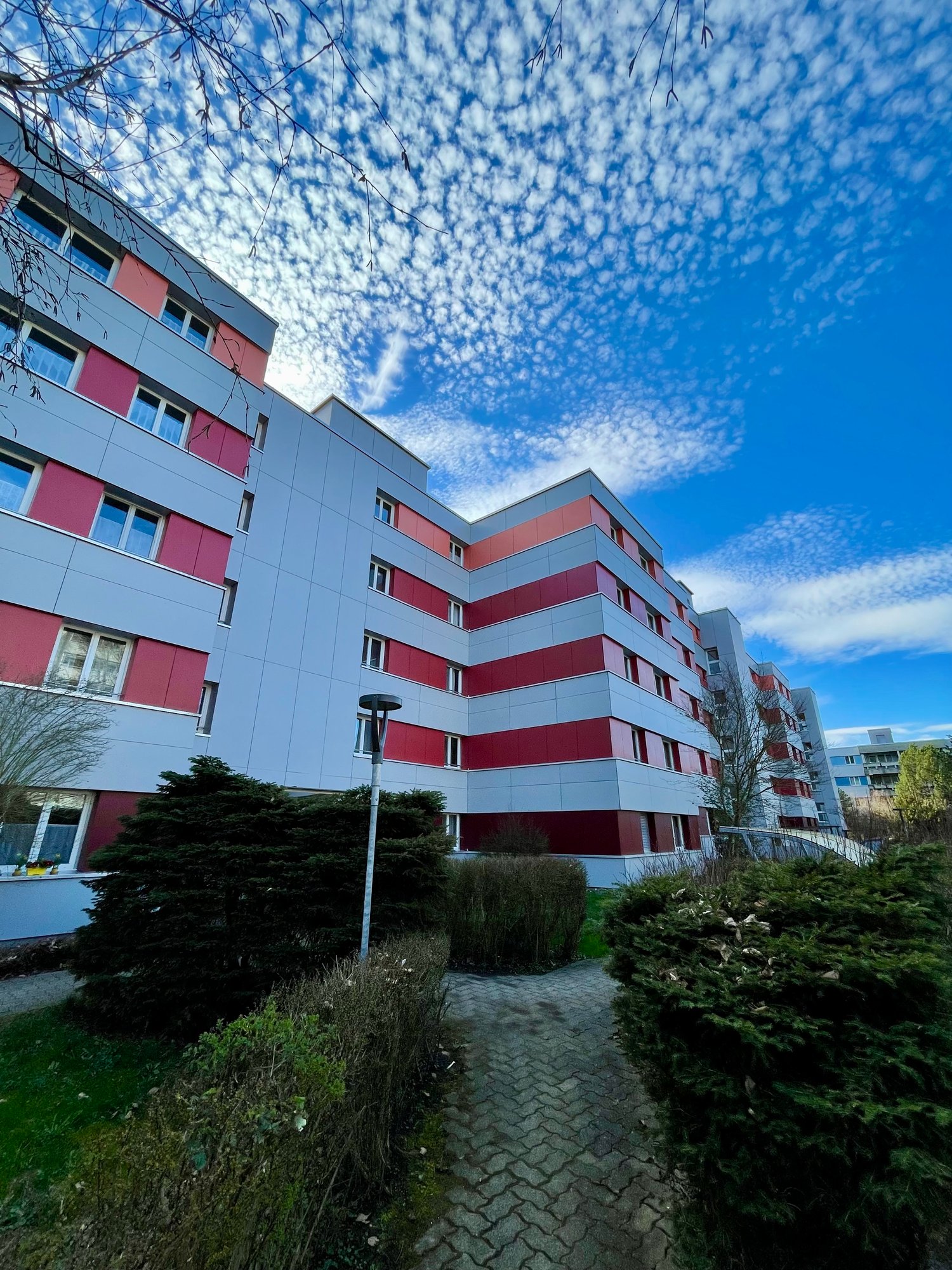 Modern apartment building with red and white exterior, multiple floors, windows, and a walkway in front.