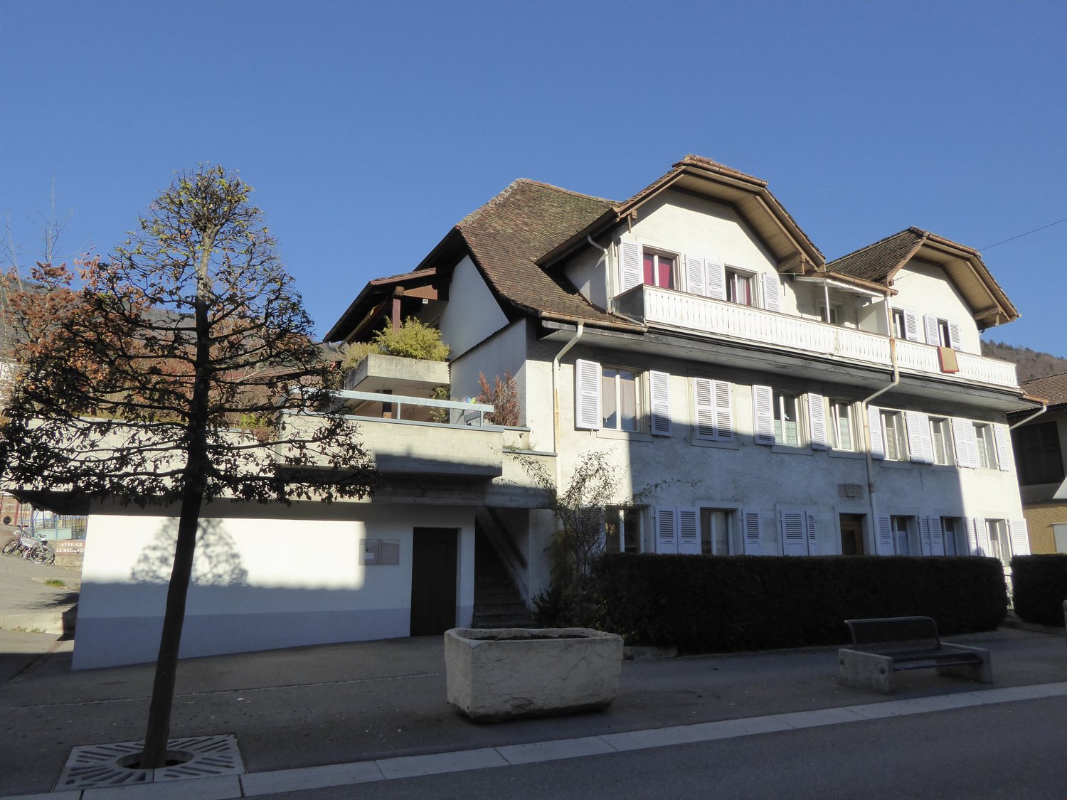 Multi-story residential building, white walls, brown roof, multiple balconies, ground floor entrance, staircase, hedge, large tree, small bench