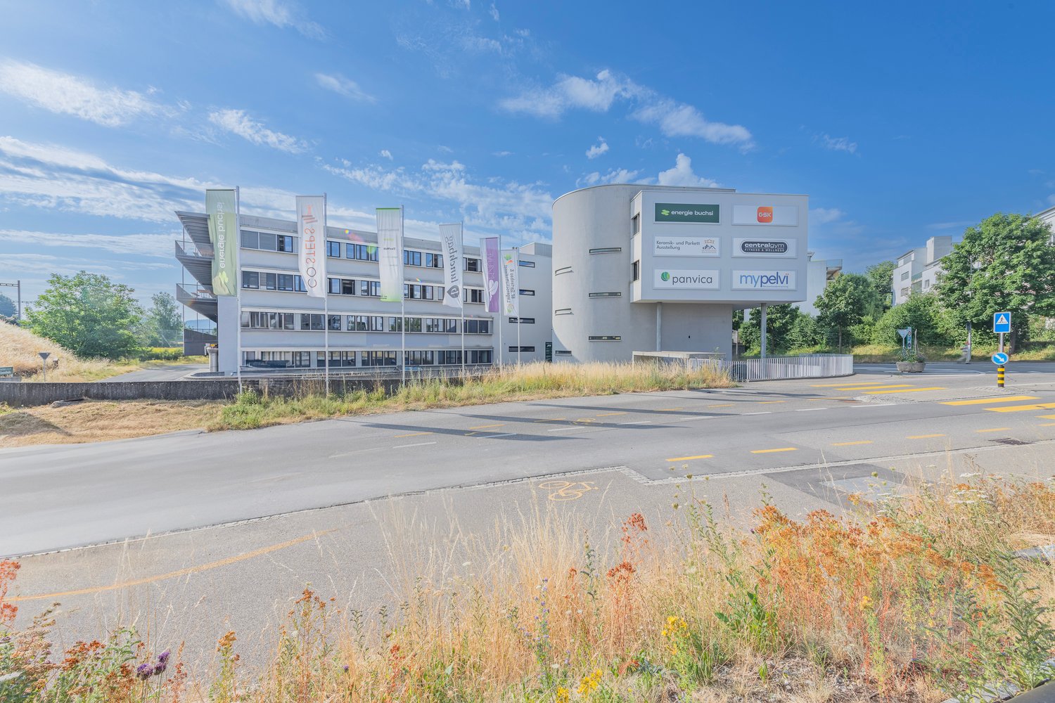 A multi-story commercial building with various company logos displayed on the facade. The building has a modern, angular design with large windows. There is a parking area in front of the building and some vegetation and grass surrounding it.