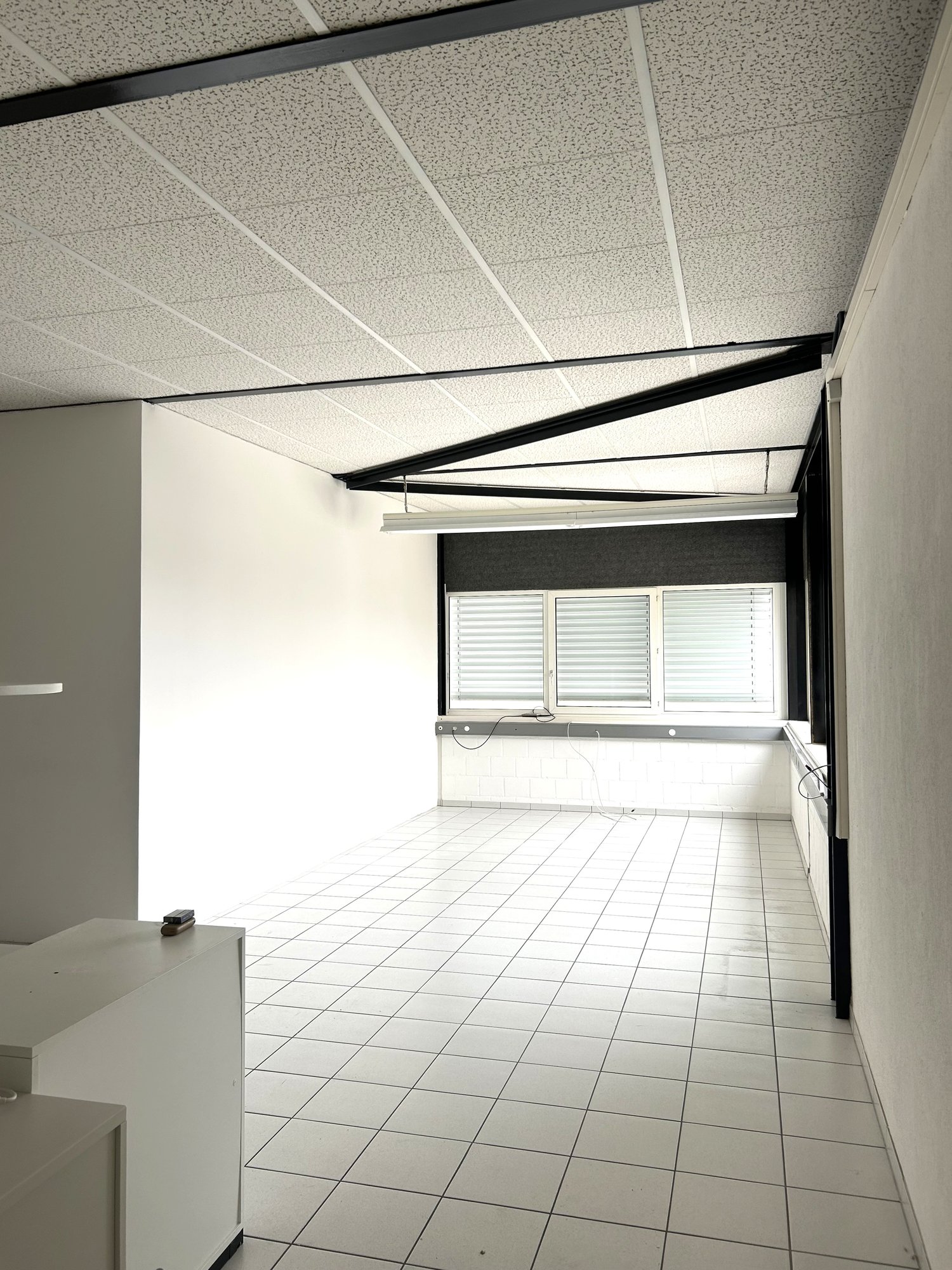 Empty office room with white tiles, modern ceiling with lighting, white walls, window with blinds, and white cabinet in the corner