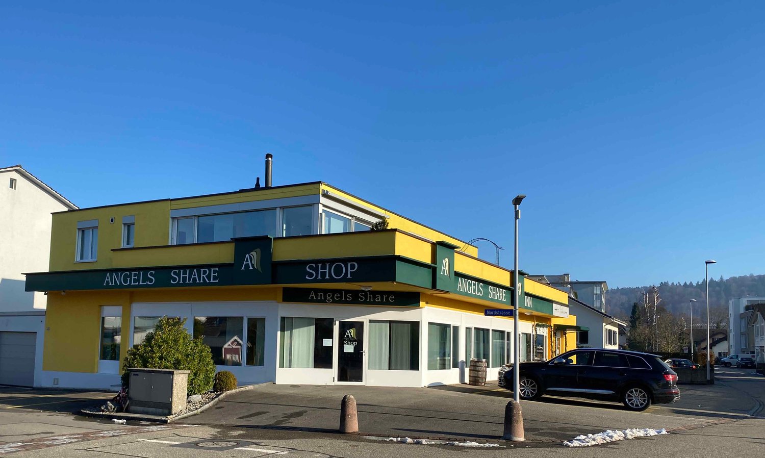 Commercial building with yellow facade, glass windows, name 'Angels Share', entrance door, a car parked in front, and streetlights on the side.