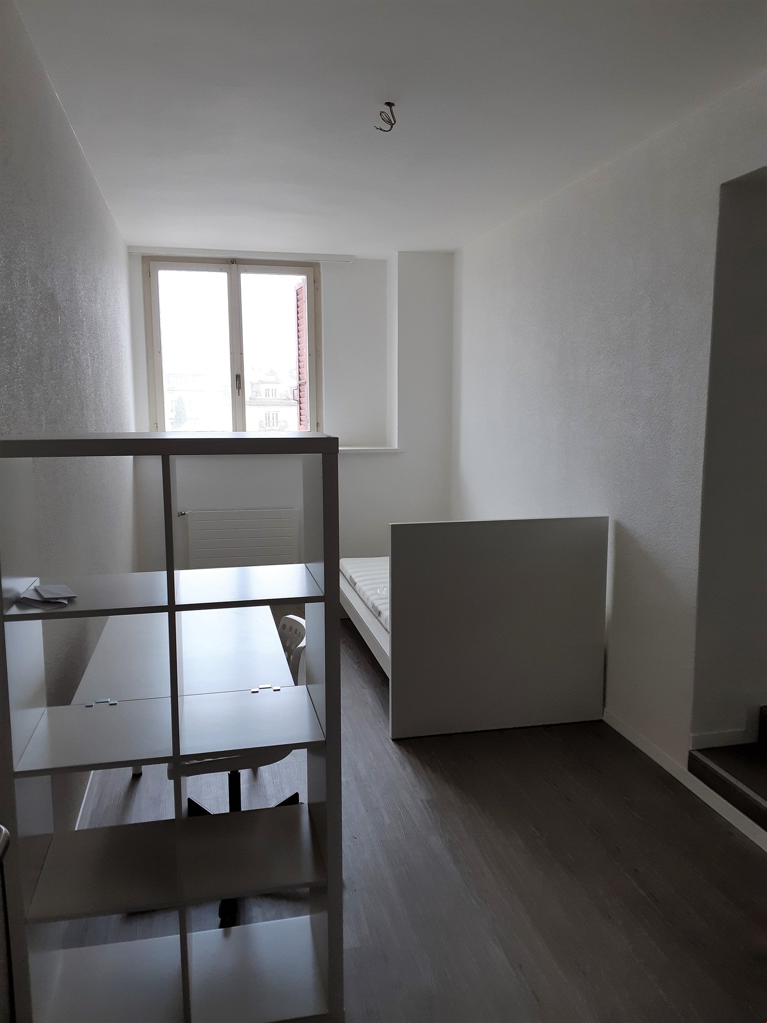 Minimalist bedroom with white walls, hardwood flooring, a metal shelving unit, and a bed frame. The room appears to be in a state of renovation or furnishing.