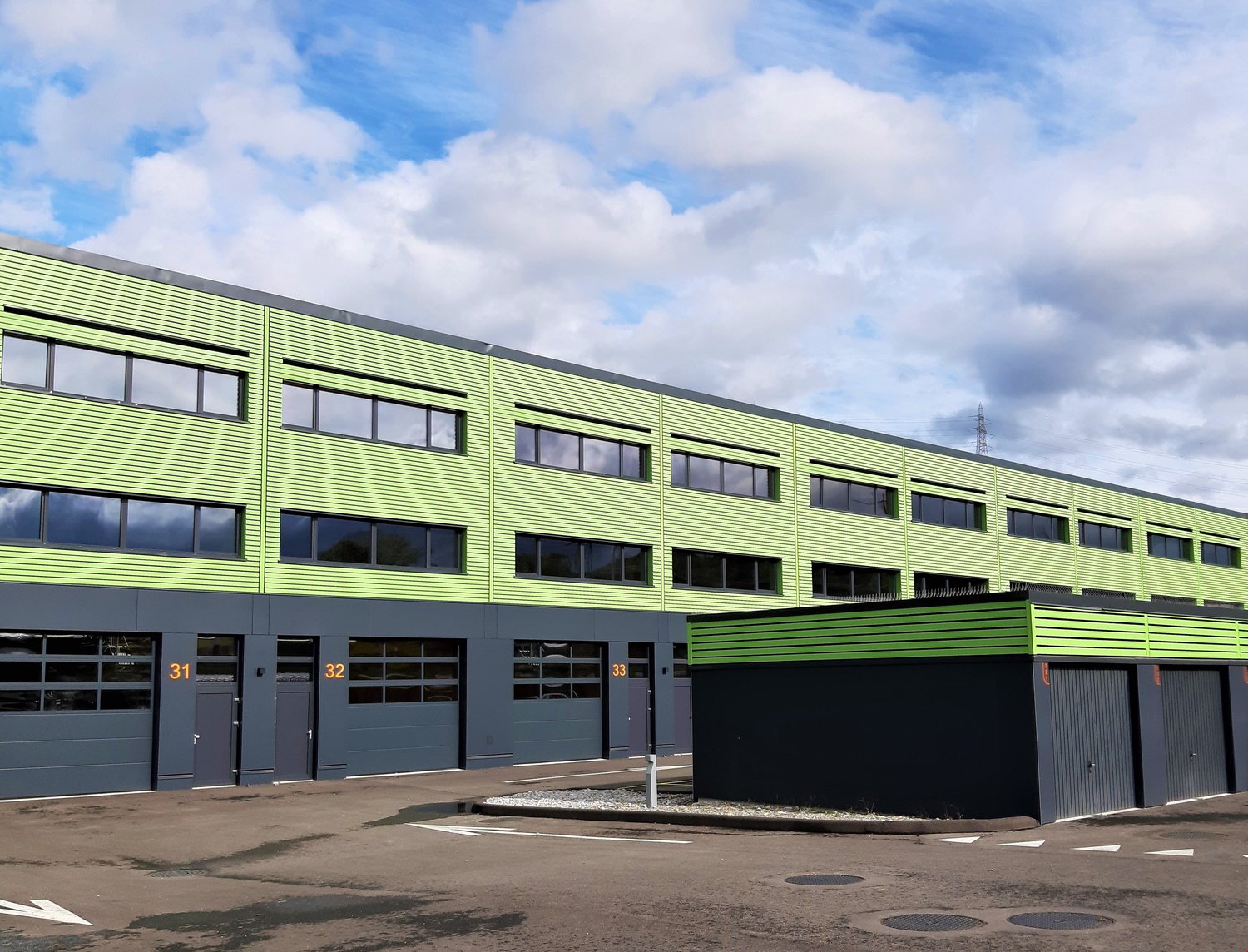 Large green and grey industrial building, several large windows, roll up garage doors on the ground floor, three garages on the side