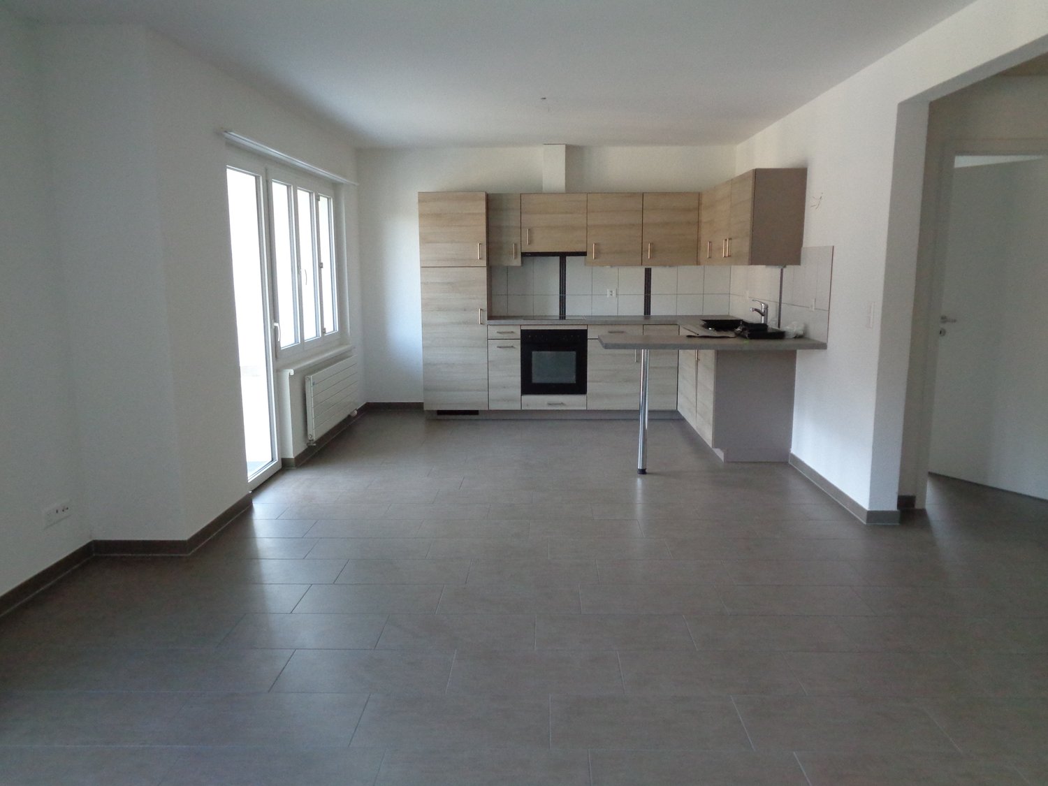 Empty kitchen with tiled floor, wooden cabinets, white walls, large windows, and a countertop with a sink and faucet.