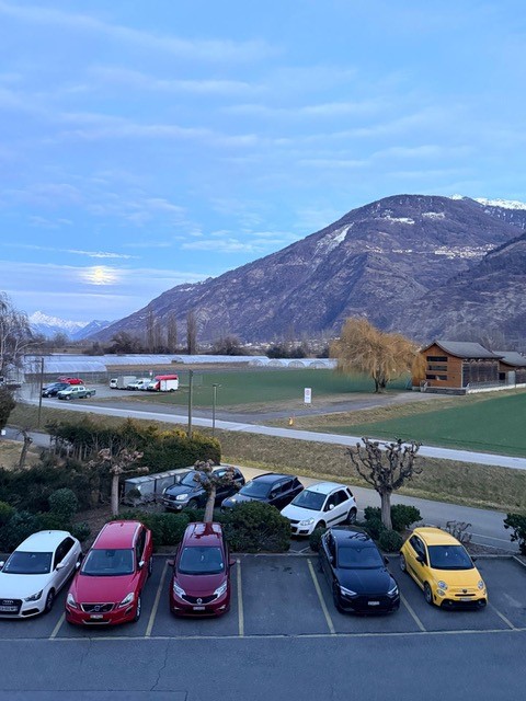 Parking lot with multiple cars parked, mountains in the background, and a building on the right side