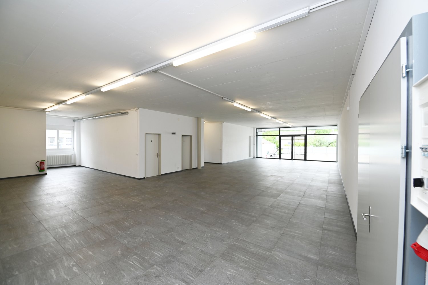Wide open space, concrete floor, two doors, fire extinguisher, glass walls, white walls, ceiling lights