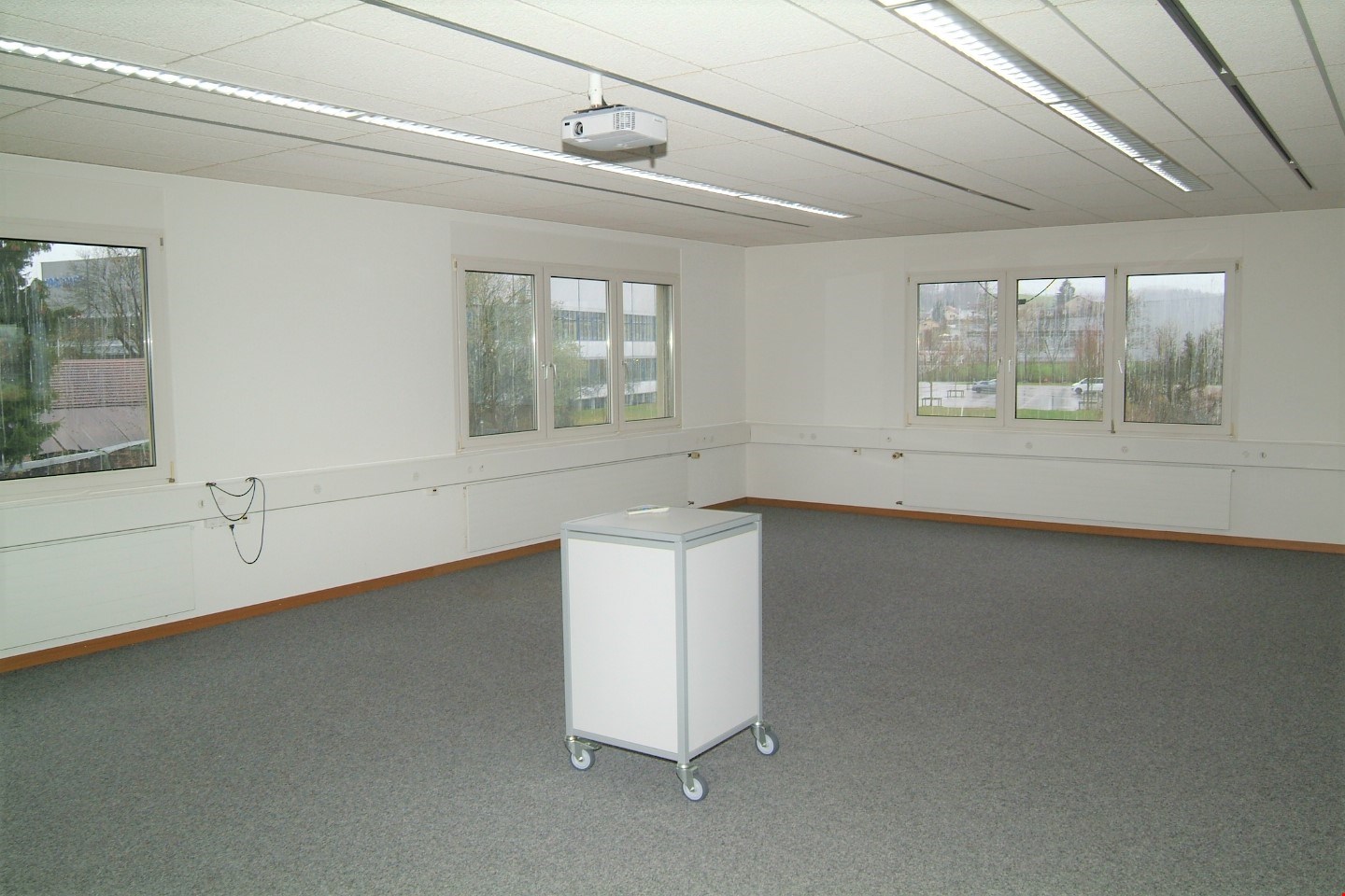 Large open room with white walls, ceiling-mounted projector, and multiple windows providing natural light. The room has a carpeted floor and a mobile cart or cabinet in the center.