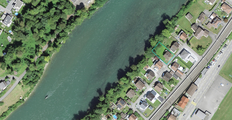 This image shows an aerial view of a residential area along a river or stream. The area is surrounded by trees and greenery, with a mix of single-story and multi-story homes. Some of the homes appear to have balconies or terraces visible. The river or str