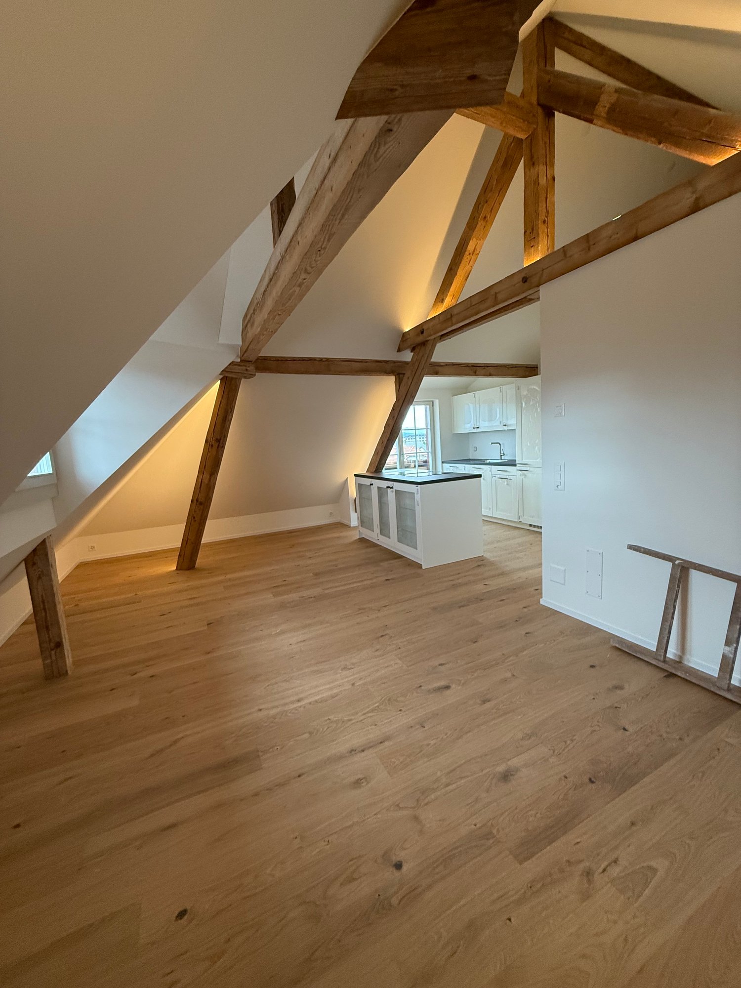 The image shows an attic or loft space with exposed wooden beams and trusses. The floor is made of hardwood planks, and there is a kitchen area visible in the background. The space has a cozy and rustic feel with the wooden elements.