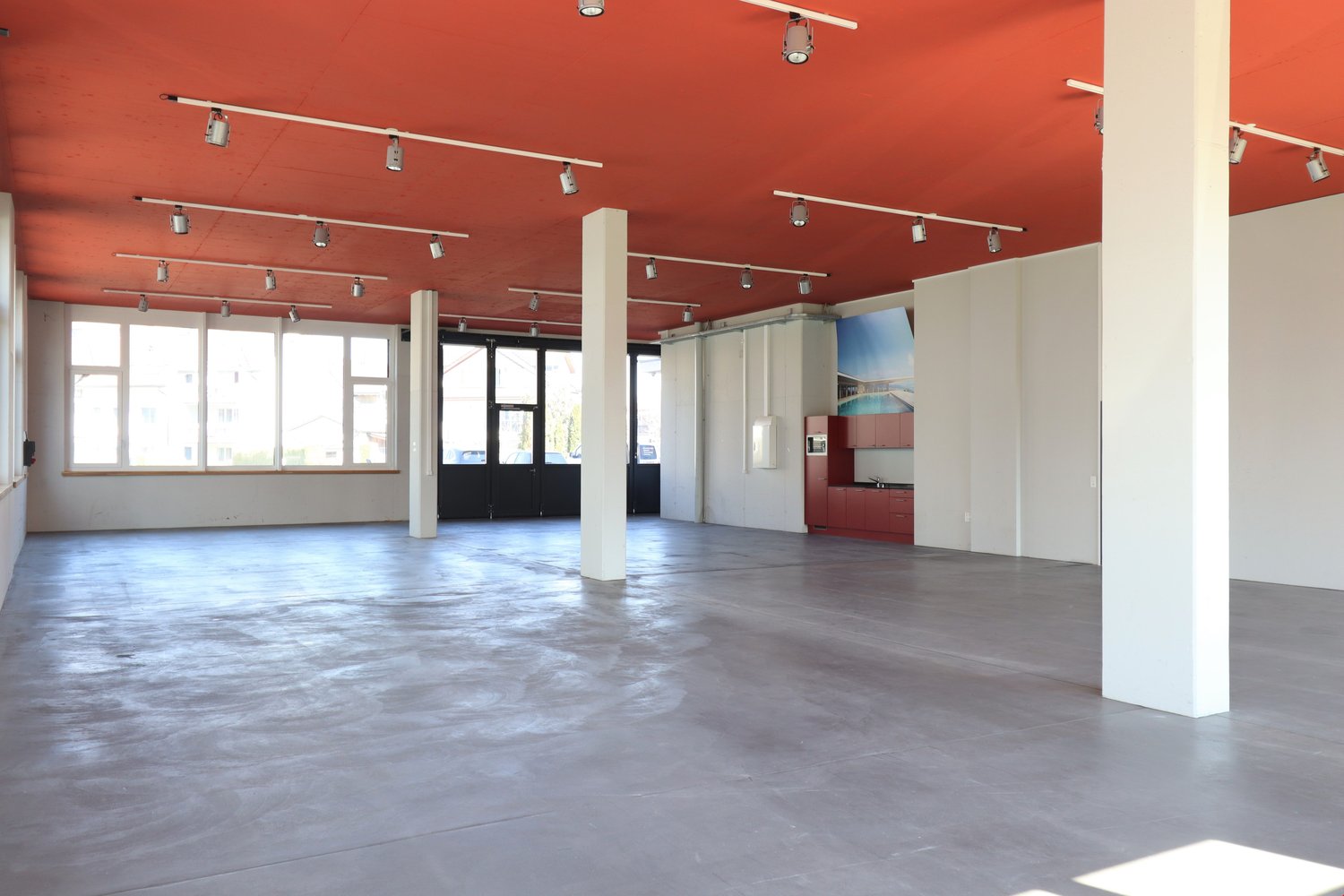 Empty room with red ceiling, white walls, concrete floor, large windows, and track lighting.