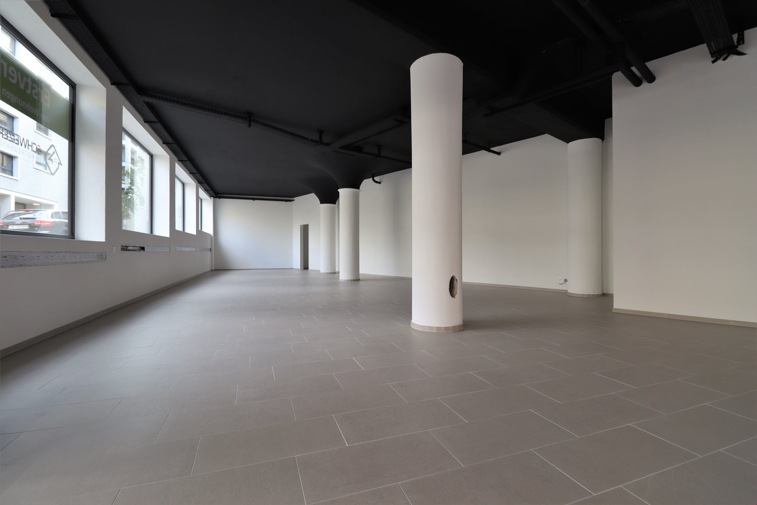 Empty room, white walls, black ceiling, gray tile floor, large windows, pillars