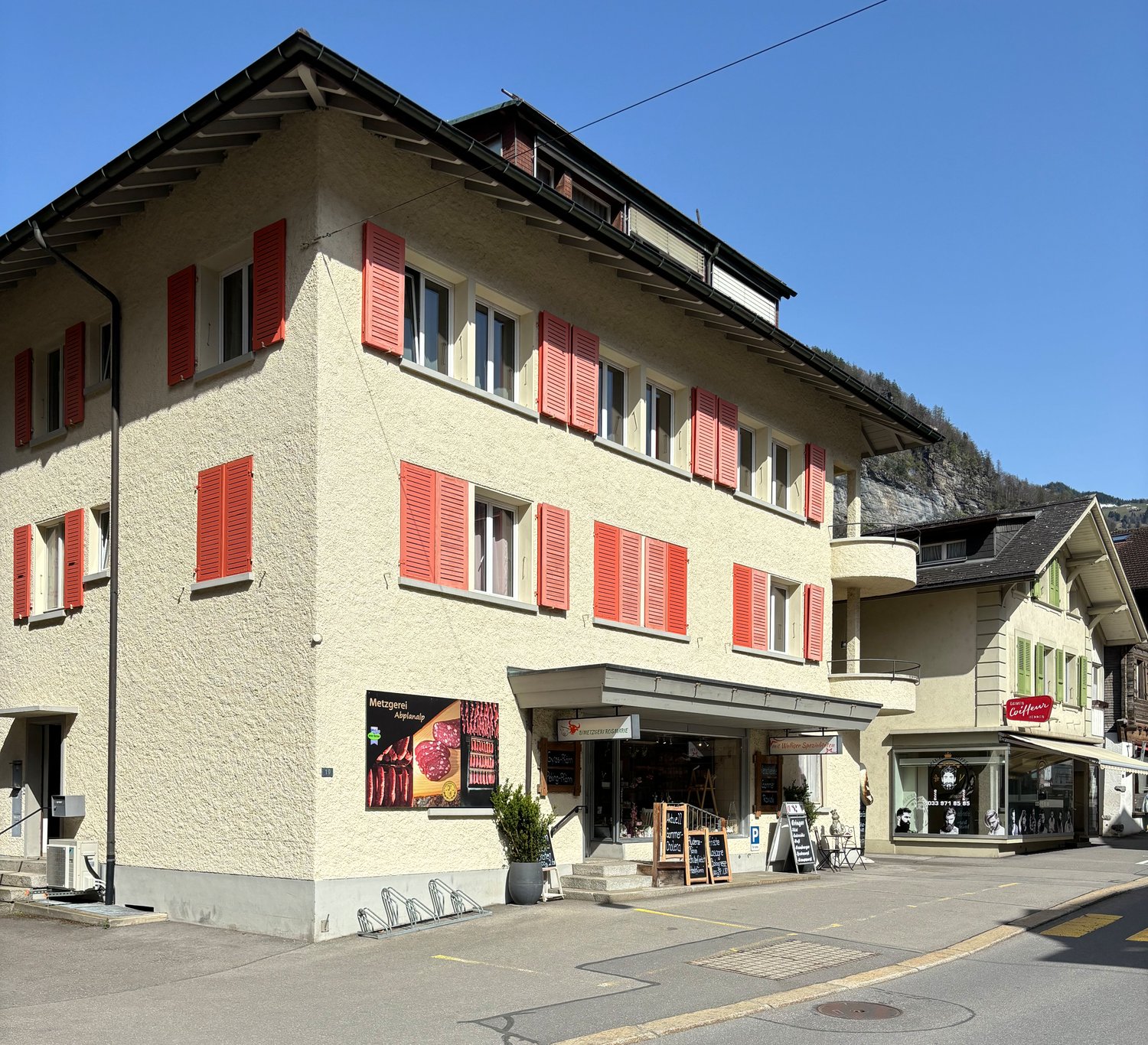 A multi-story building with a light-colored exterior and red shutters on the windows. The building has a retail shop on the ground floor and appears to be located in a small town or village setting.