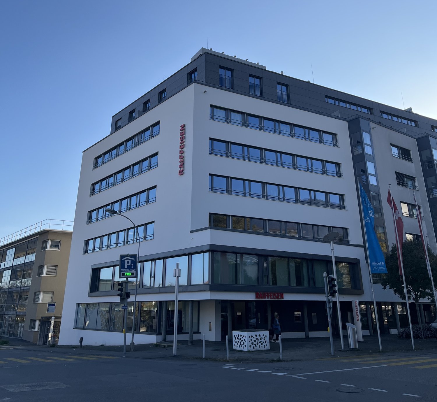 White building with 6 floors, modern design, large windows, glass doors, flags on the right side, street signs, parking sign, traffic light