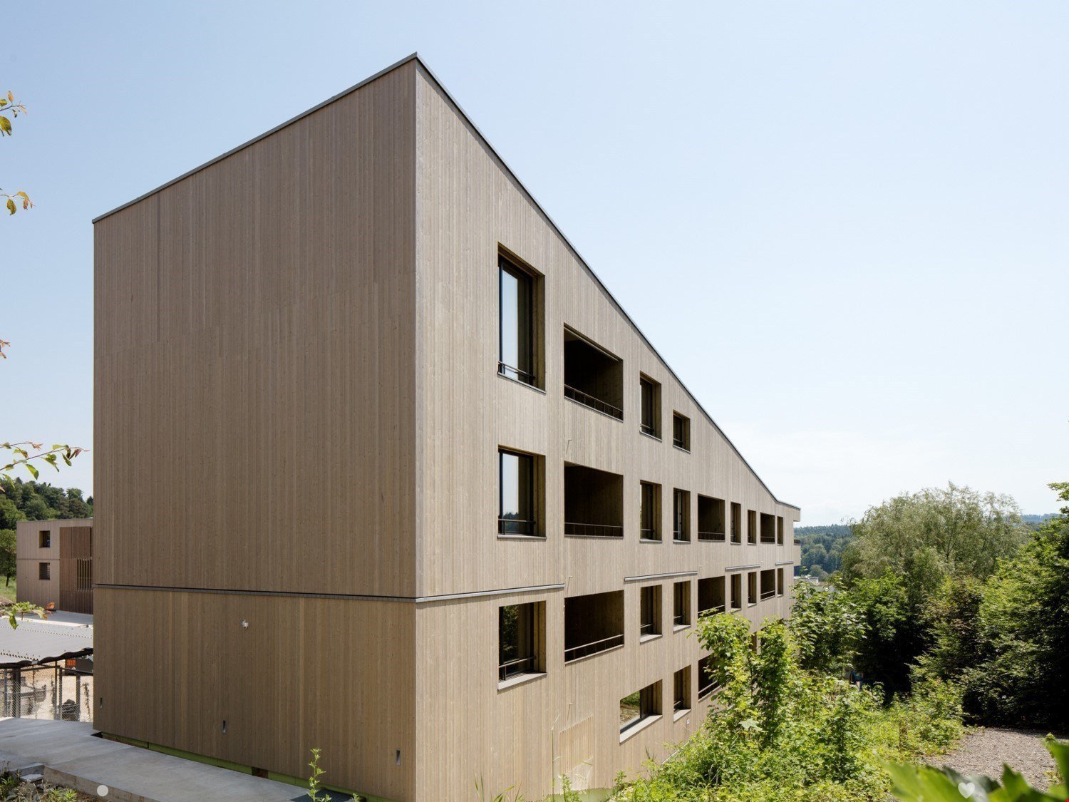 Modern wooden building, multiple windows, balconies, surrounded by greenery