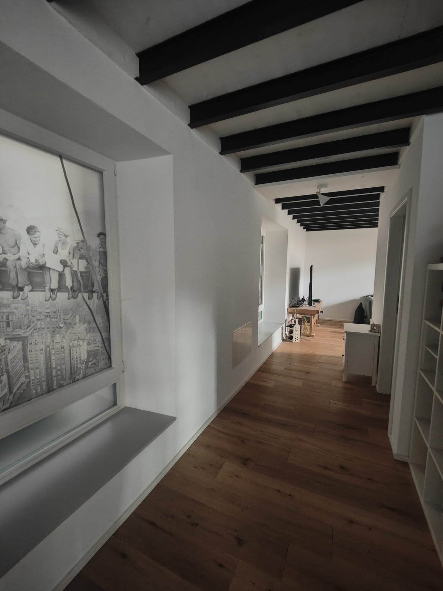 Empty hallway with wooden floor, white walls, black beams, large window with a picture, small table with TV, white shelves