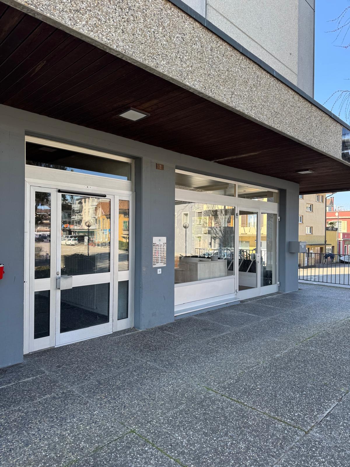 Exterior of a building with glass doors and windows, concrete floor, metal railings, and a small signboard.
