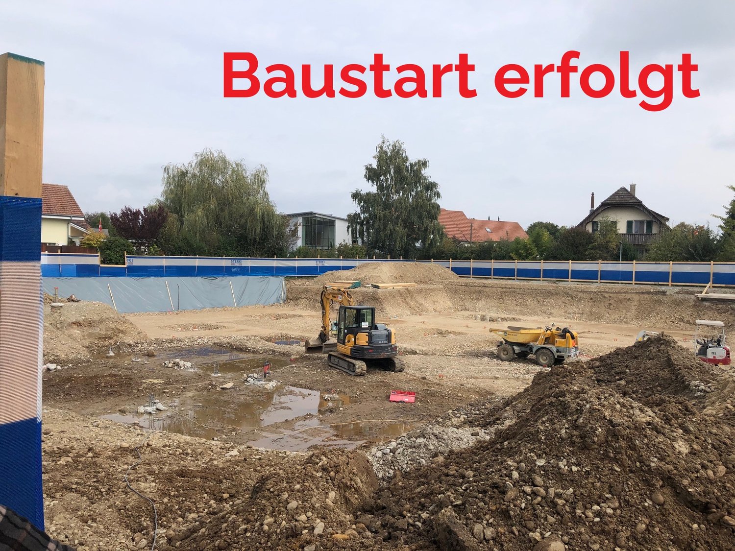 Construction site, multiple vehicles, dirt, temporary fence, houses in background