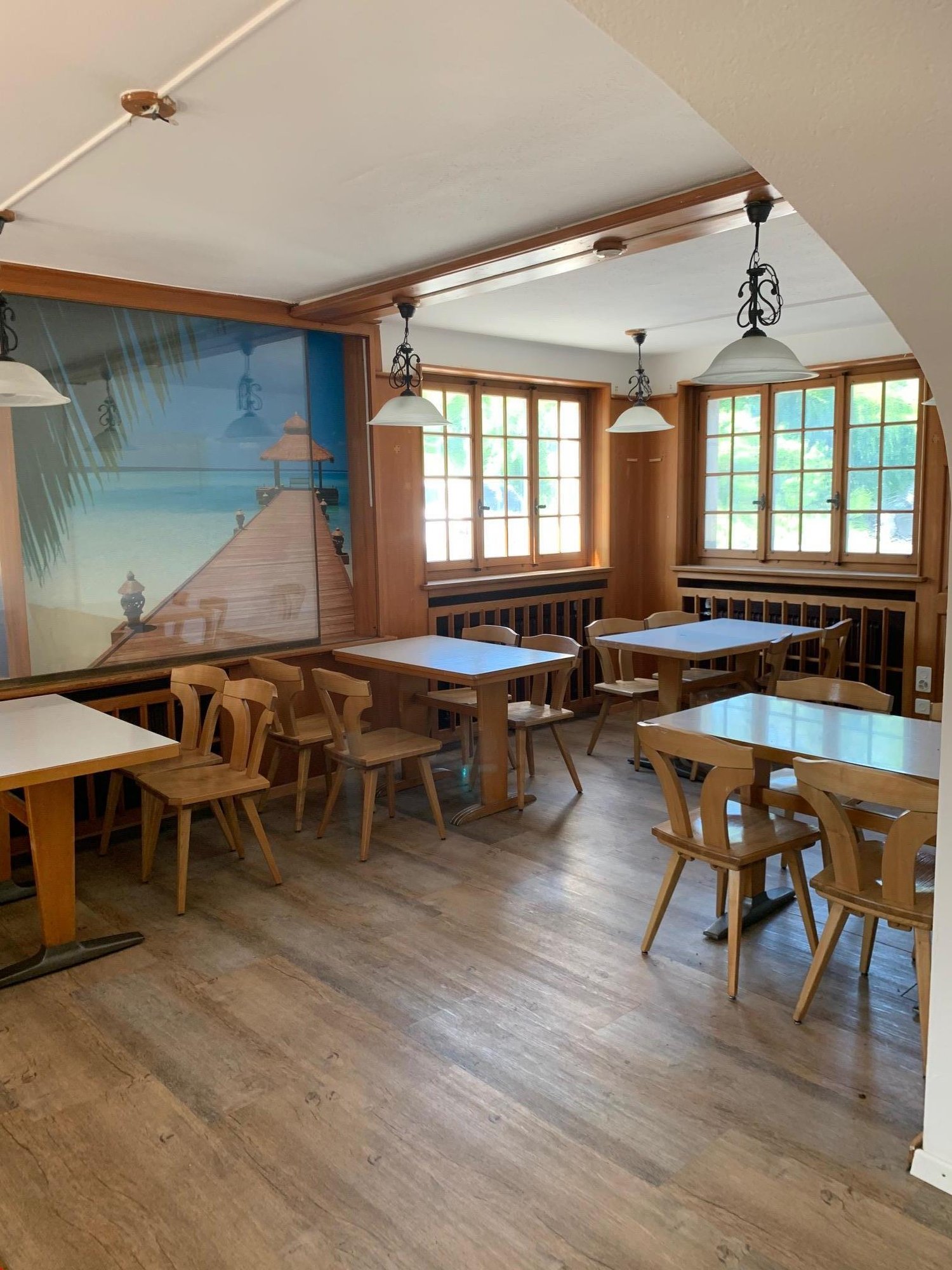 Cozy dining room with wooden tables and chairs, large windows, and a mural depicting a beach scene on the wall.