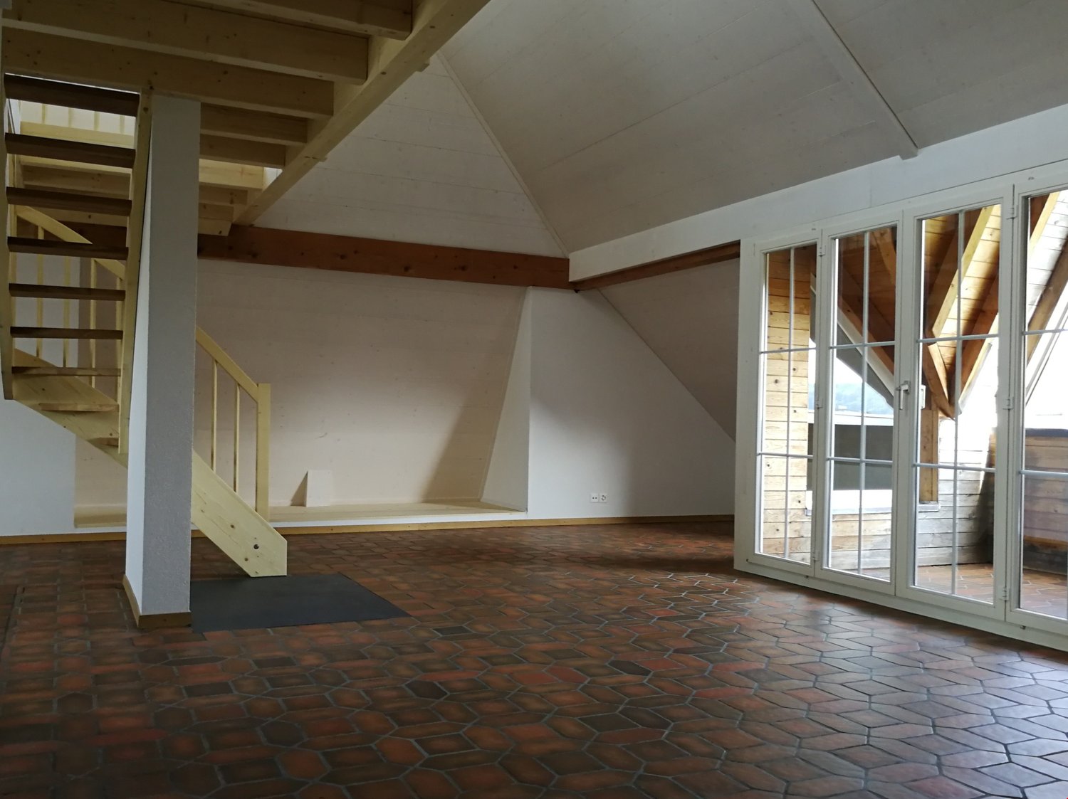 Empty room with wooden staircase, white walls, wooden beams, and red hexagonal tiles on the floor.