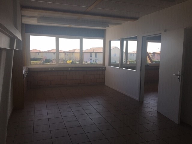 Large open space with tile flooring, multiple windows providing natural light, and a balcony or terrace visible through the windows.