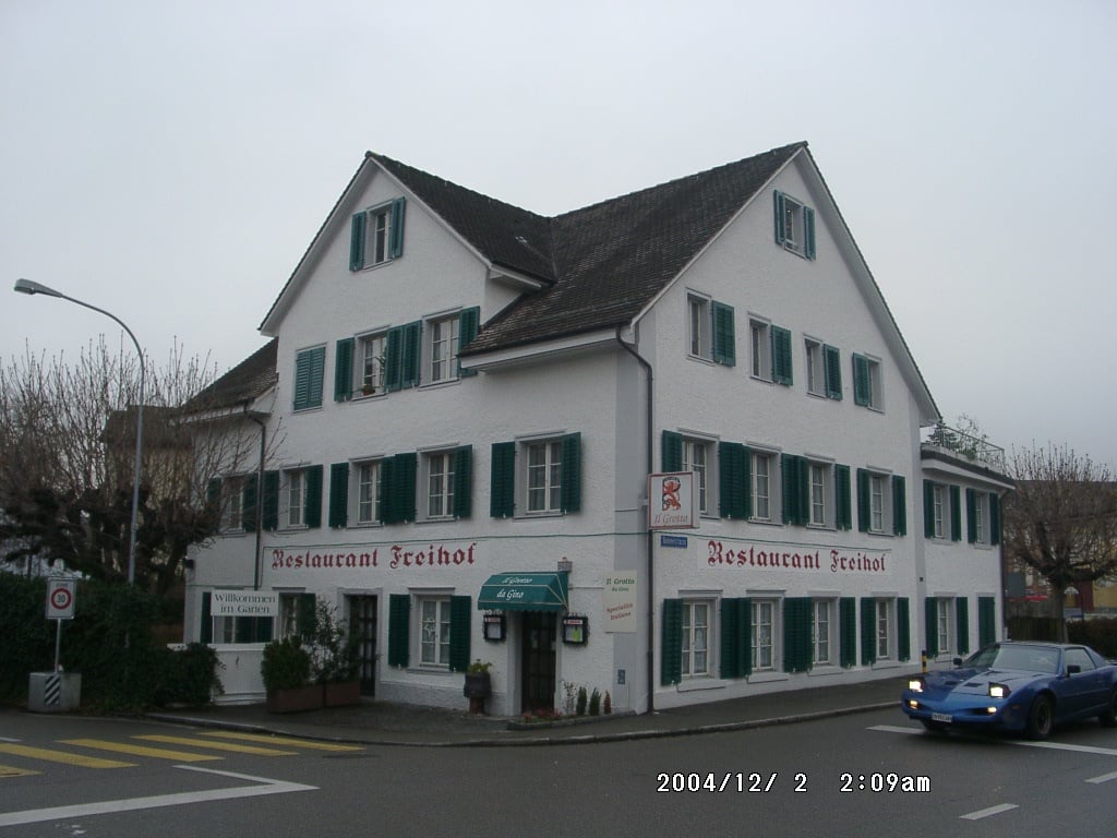 2-story restaurant building, white exterior, green shutters, red text sign, shrubs and plants in front, streetlight and blue car in the background